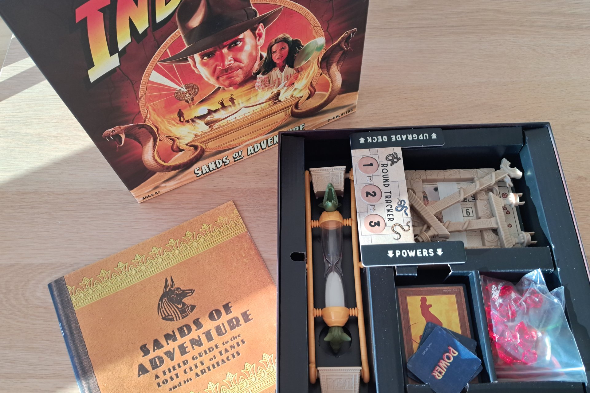 The opened box of the Indiana Jones Sands of Adventure board game, showcasing its components. The box contains various items such as game cards, a game board, player tokens, dice, and small plastic figures representing Indiana Jones and other characters.